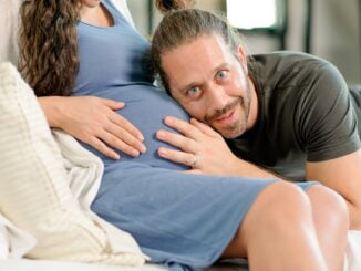 Man sit and listen baby sound from his pregnancy wife belly and he look happy also look to camera