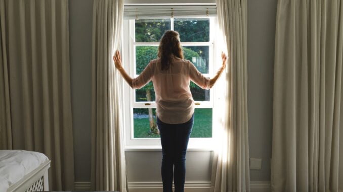 Thoughtful senior caucasian woman in bedroom, standing next to window, opening curtains
