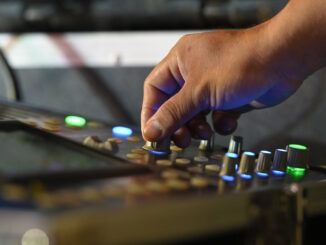 Close-up of a sound engineer's hand adjusting knobs on an audio mixers console, with illuminated buttons and sliders indicating levels, showcasing the intricate process of audio mixing.