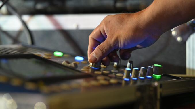 Close-up of a sound engineer's hand adjusting knobs on an audio mixers console, with illuminated buttons and sliders indicating levels, showcasing the intricate process of audio mixing.